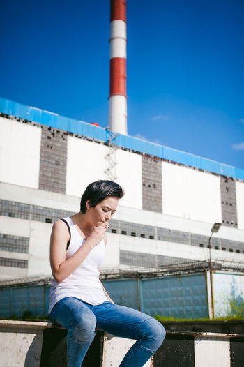 Young woman smoking cigarette while sitting on barricade against factory