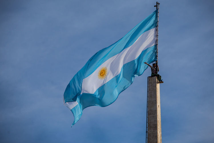 Low angle view of woman sitting on flag pole against blue sky