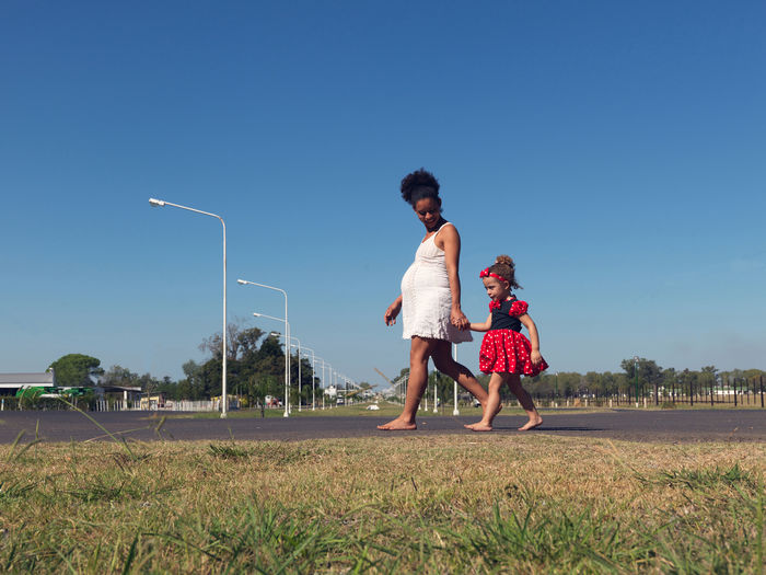 Mother and daughter walking on grassy field against clear sky