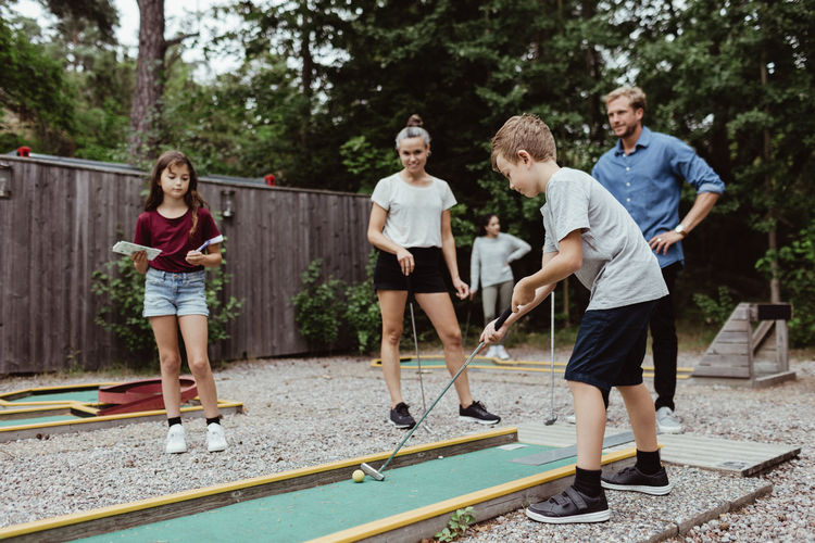 Full length of family looking at boy playing miniature golf in backyard