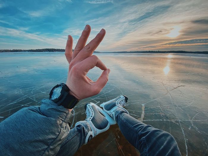 Low section of man sitting on frozen lake gesturing against sky