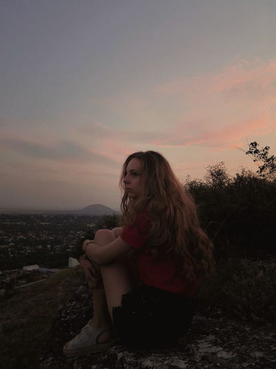 Beautiful young woman sitting in park against sky during sunset