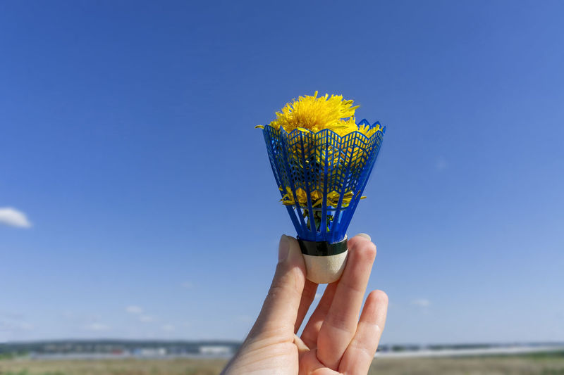Blue shuttlecock from badminton with yellow dandelions in hand against blue sky, summer background 