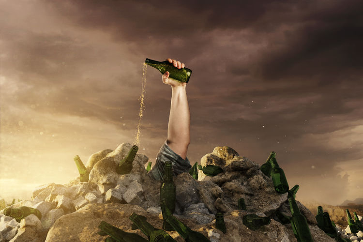 Digital composite image of cropped hand holding beer bottle against sky during sunset