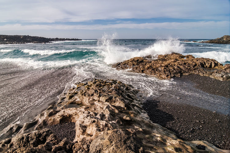 Waves splashing on rocks at shore against sky