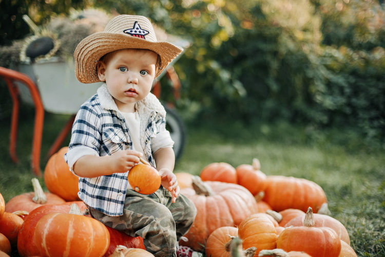 A little cowboy boy in a hat and a plaid shirt is sitting on a pile of pumpkins in the garden 