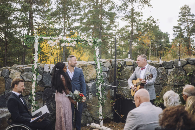 Mature man playing guitar for newlywed couple by guests and minister during wedding ceremony