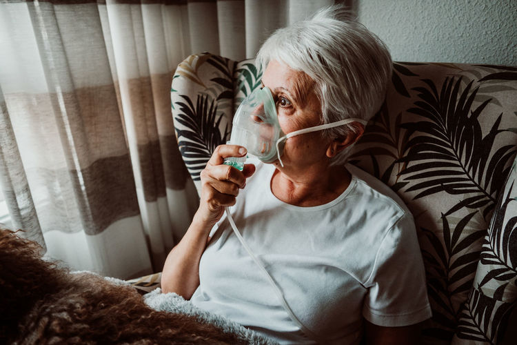 Senior woman wearing oxygen mask sitting with dog on chair at home