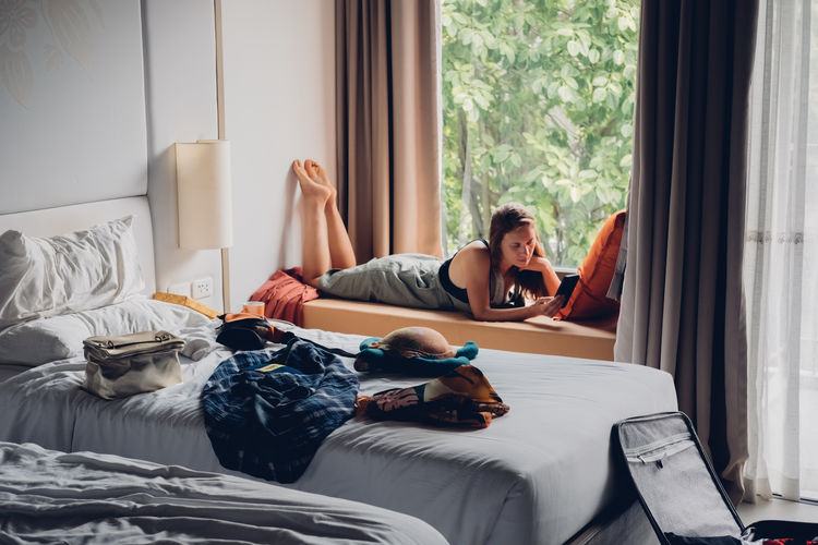 Girl student lying by the window and talking on mobile phone in room with mess.