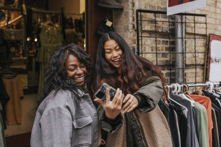 Happy female friends taking selfie through smart phone while enjoying outside clothing store