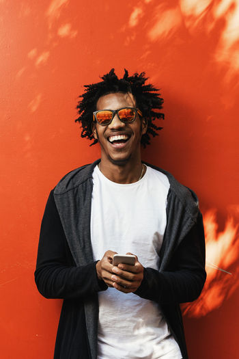 Smiling man with mobile phone standing against orange wall