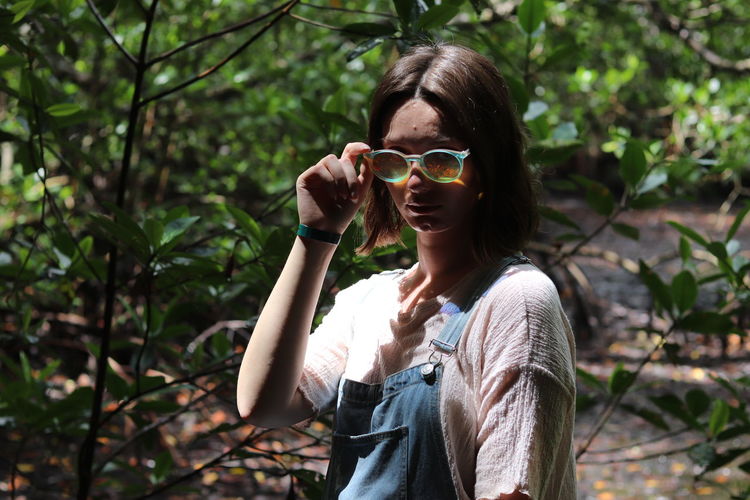 Portrait of young woman holding sunglasses against trees