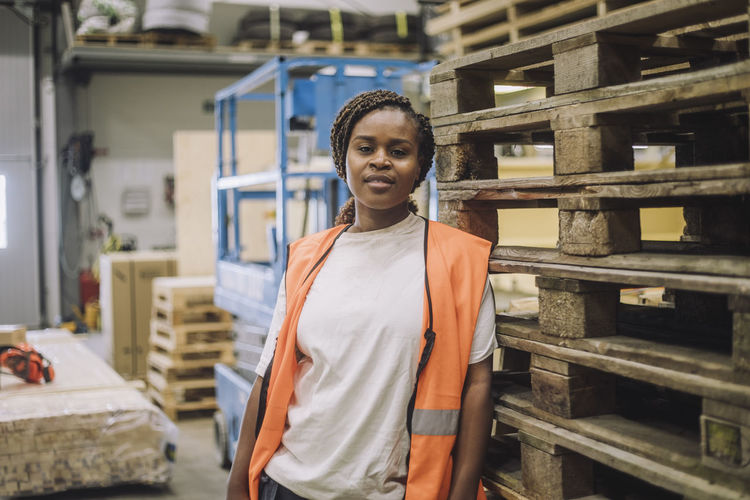 Portrait of female carpenter leaning on pallet stack in warehouse