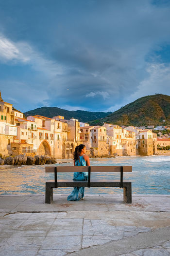 Rear view of woman sitting on bench against buildings in city