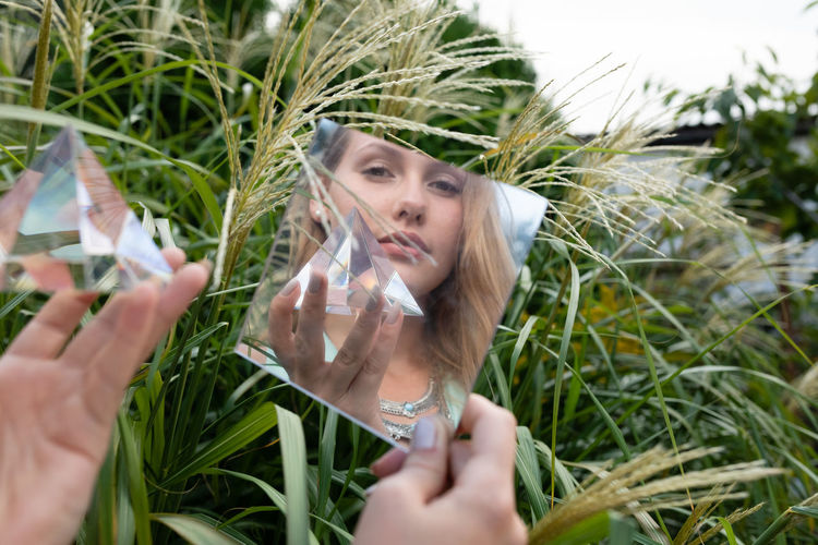 Reflection of woman looking away and holding prism in mirror against plants