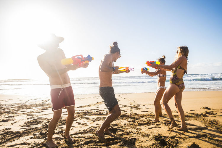 Full length of playful friends playing with squirt guns at beach during sunny day