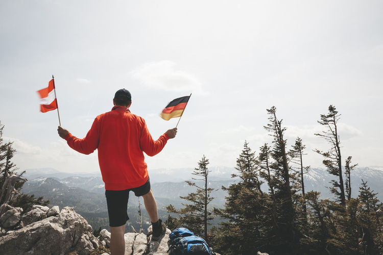 Rear view of mature man holding national flags while standing on mountain against sky