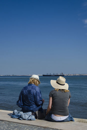 Rear view of couple sitting on retaining wall at pier against blue sky