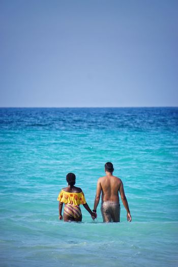 Shirtless man holding hand of woman while standing in sea against clear blue sky