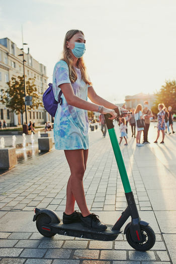 Full length of girl wearing mask standing on push scooter in city