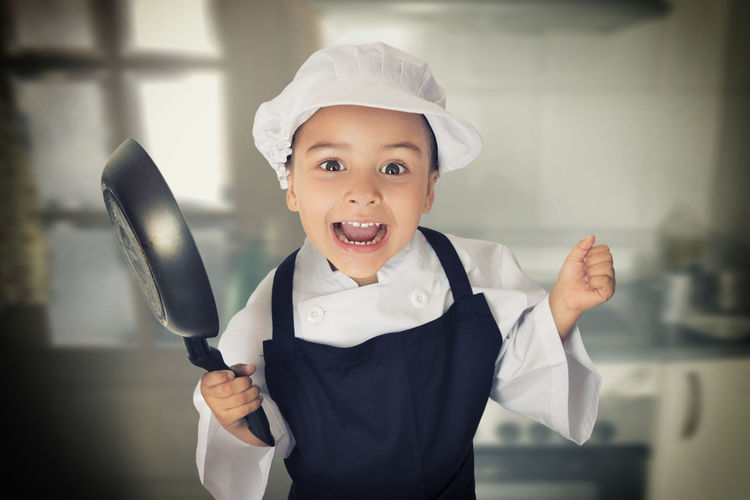 Portrait of cheerful girl wearing chef uniform holding cooking pan while standing in kitchen