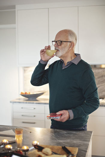 Senior man drinking juice while standing by kitchen island at home