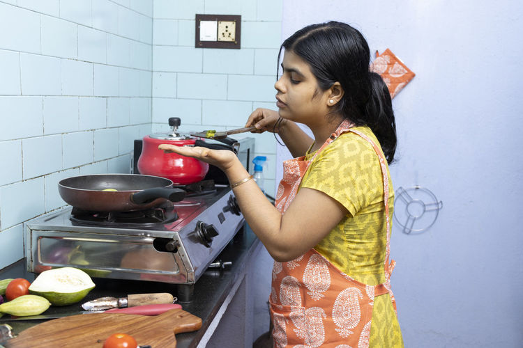 A pretty indian young woman wearing apron cooking and tasting food in domestic kitchen on gas stove