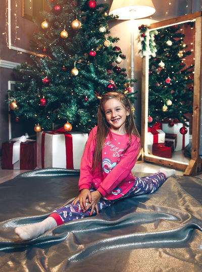 Full length of happy girl stretching legs while sitting against christmas tree at home