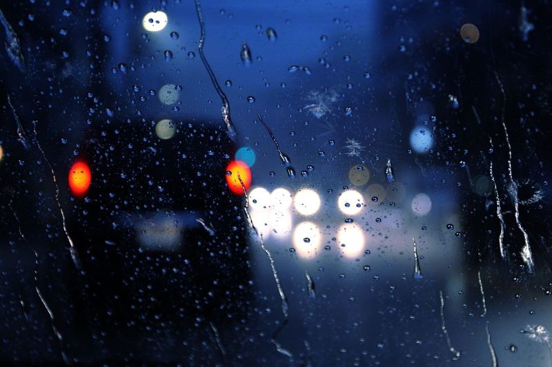 Illuminated cars on city street seen through wet glass window during monsoon