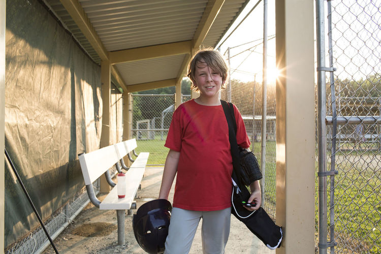 Portrait of smiling boy with sports equipment standing in baseball dugout at playing field