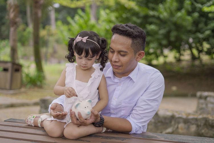 Smiling father and daughter inserting coin in piggy bank while sitting outdoors