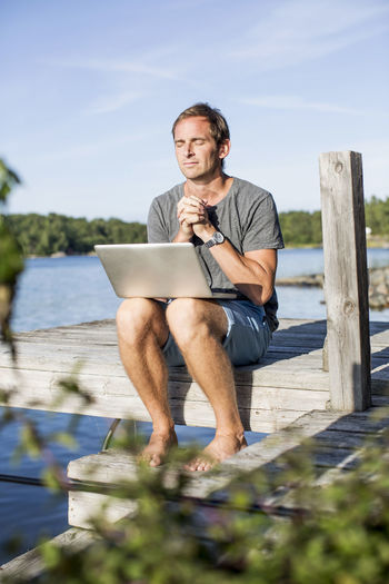 Mature man praying while sitting with laptop on pier