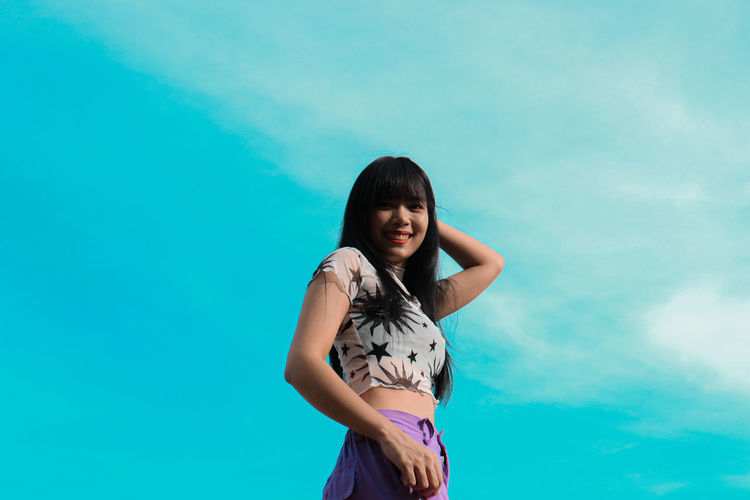 Low angle portrait of young woman standing against blue sky