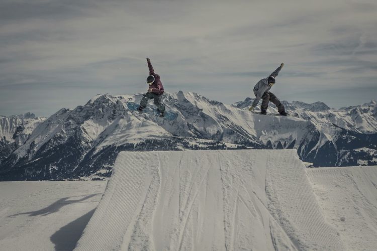 Snowboarders jumping over snow covered ramp at mountains against sky