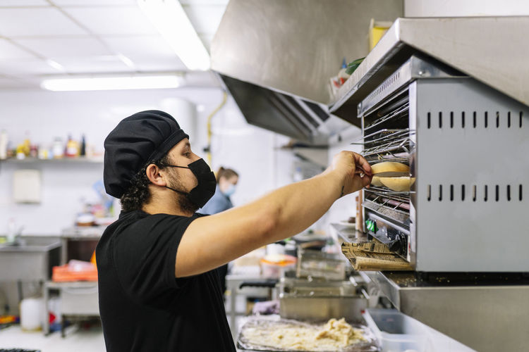 Male chef preparing bread in oven at restaurant kitchen during covid-19
