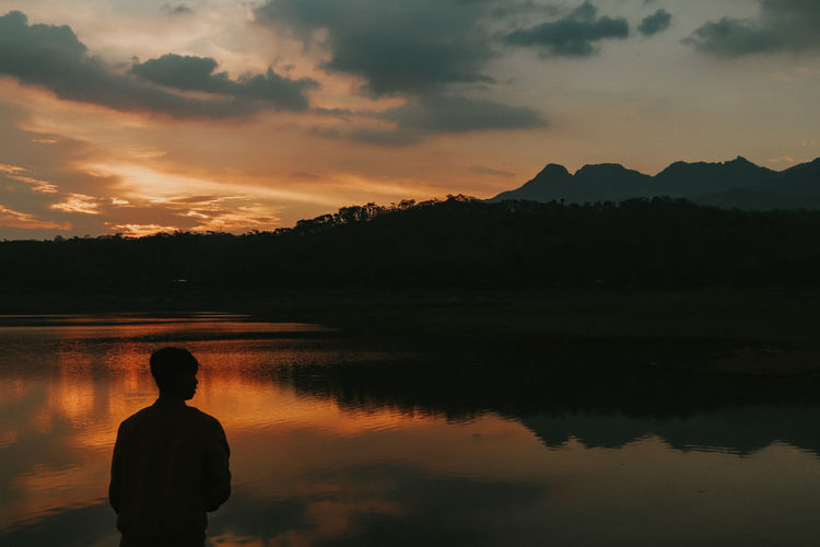 Silhouette man looking at lake against sky during sunset