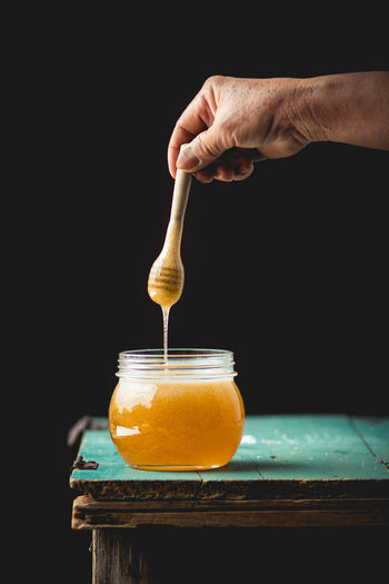 Cropped hand holding honey dipper over glass jar on table against black background