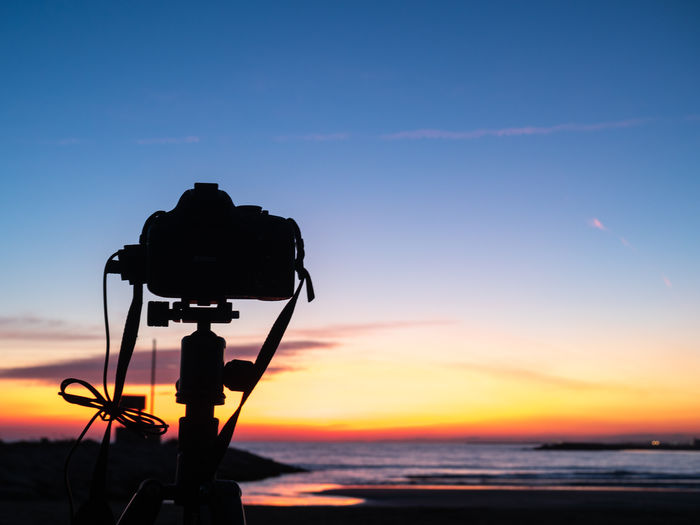 Silhouette of camera by sea against sky during sunset