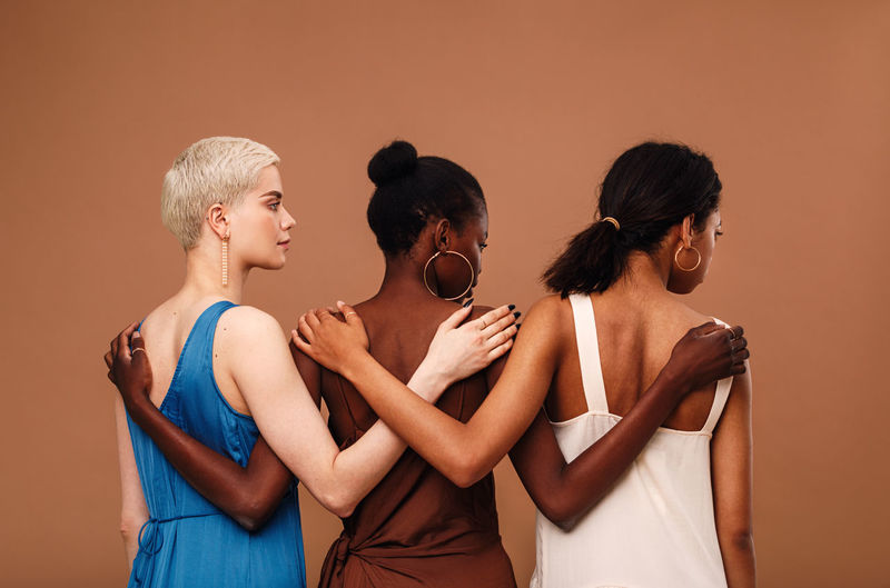 Multi-ethnic women with arm around standing against brown background