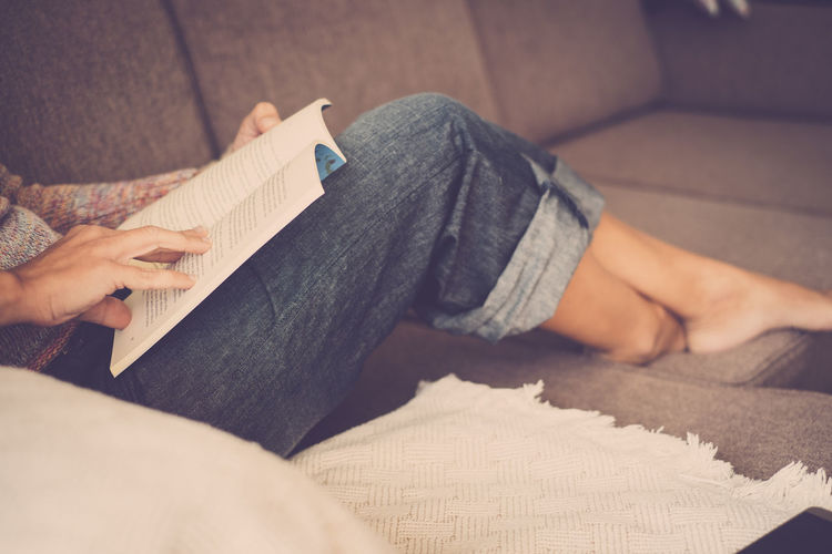 Low section of woman reading book on sofa at home