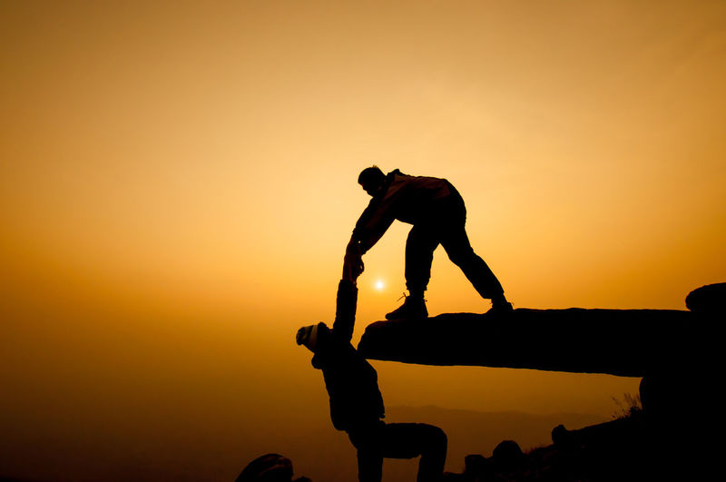 Silhouette man assisting friend climbing cliff against orange sky during sunset
