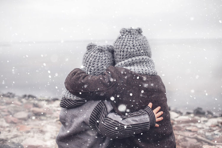 Rear view of sibling embracing while standing by lake during winter