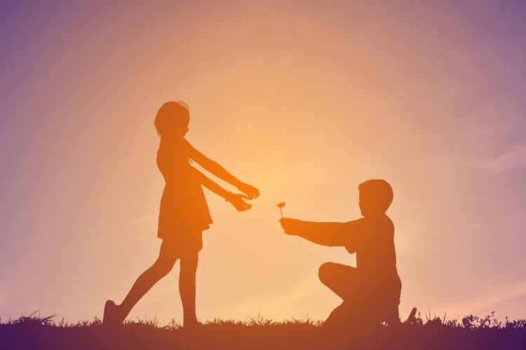 Low angle view of teenage boy offering flower to girl on field against sky during sunset