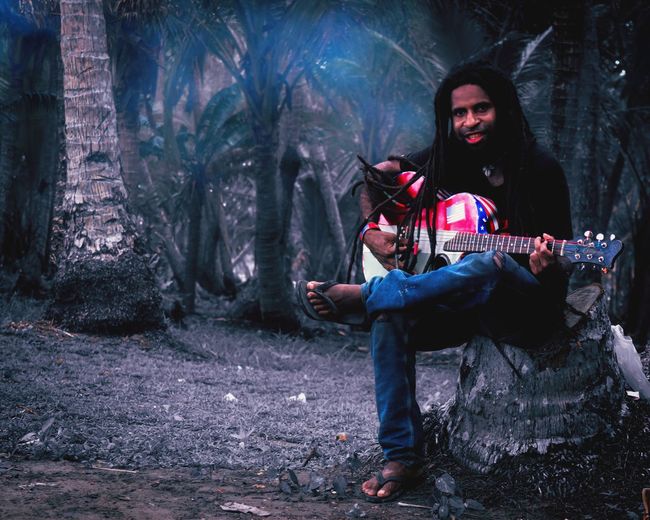 Smiling man playing guitar sitting against trees