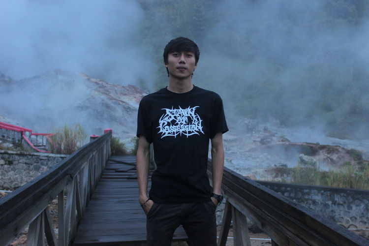 Portrait of young man standing on footbridge against mountains