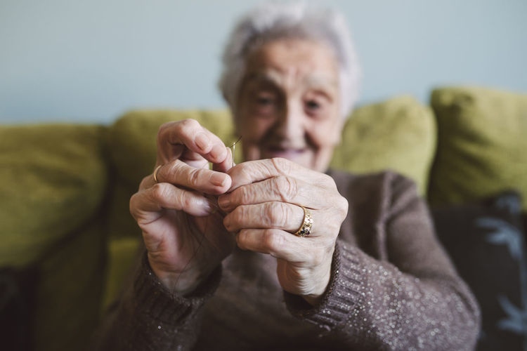 Hands of senior woman sitting on couch passing thread through buttonhole of sewing needle