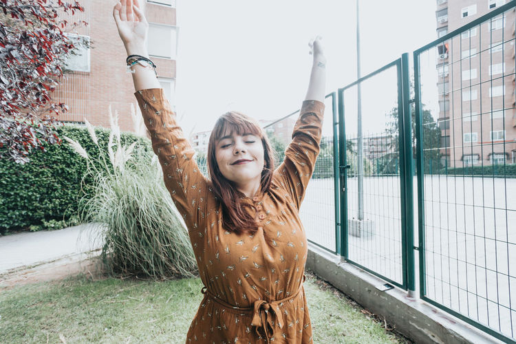 Portrait of smiling young woman with arms outstretched standing against trees