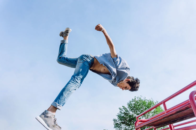 Low angle fearless young man jumping above metal railing in city while performing parkour stunt on sunny day