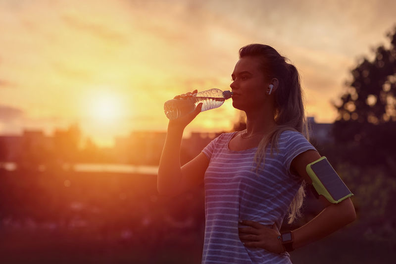 Young woman drinking glass while standing against sunset