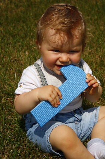 Baby boy playing with toy while sitting on grassy field at park during sunny day